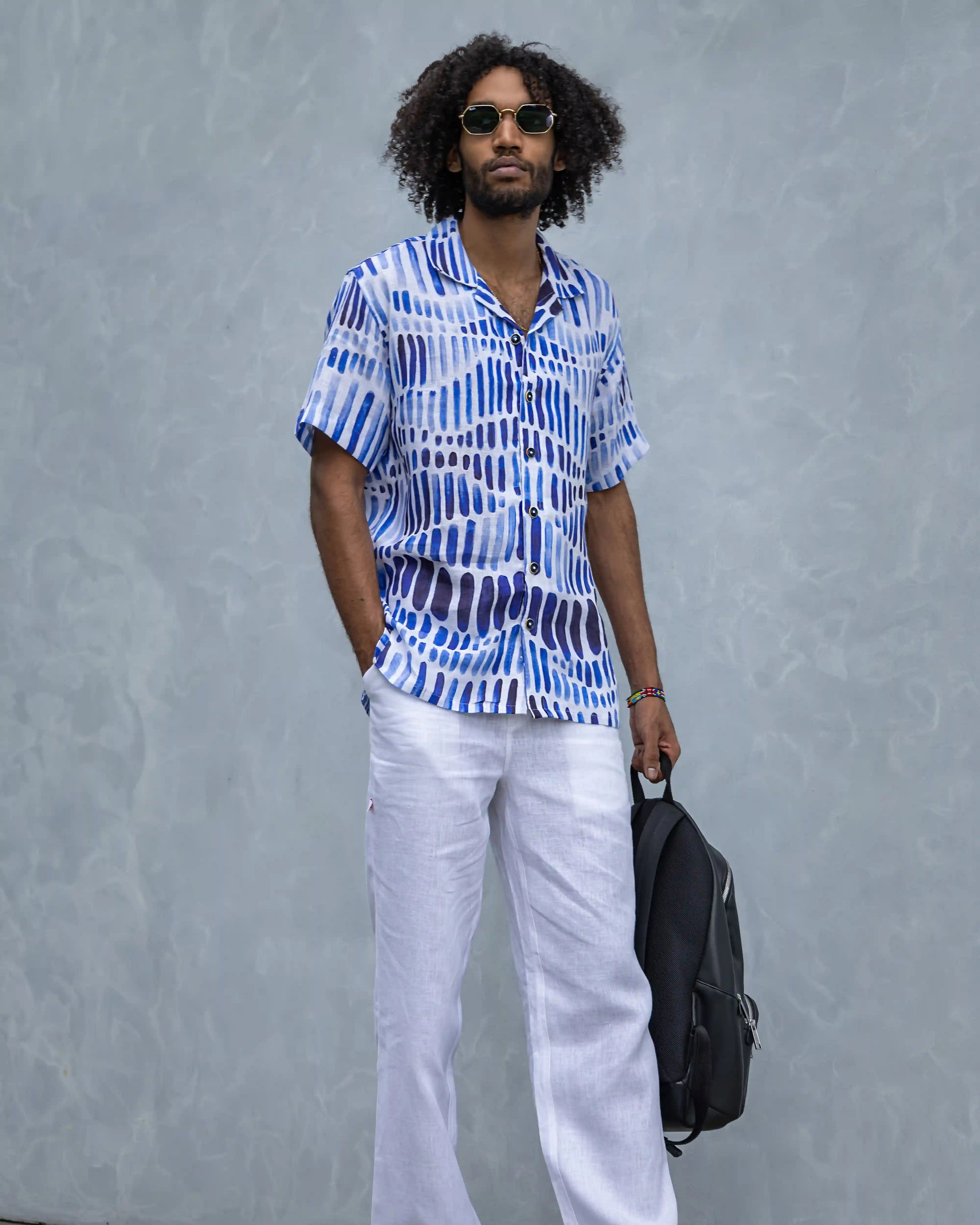 A model in a NAD's blue watercolour printed shirt for men, white linen pants, and a black bag.